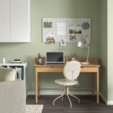Modern home office: light wooden desk, white chair, green wall; pegboard, laptop, lamp.