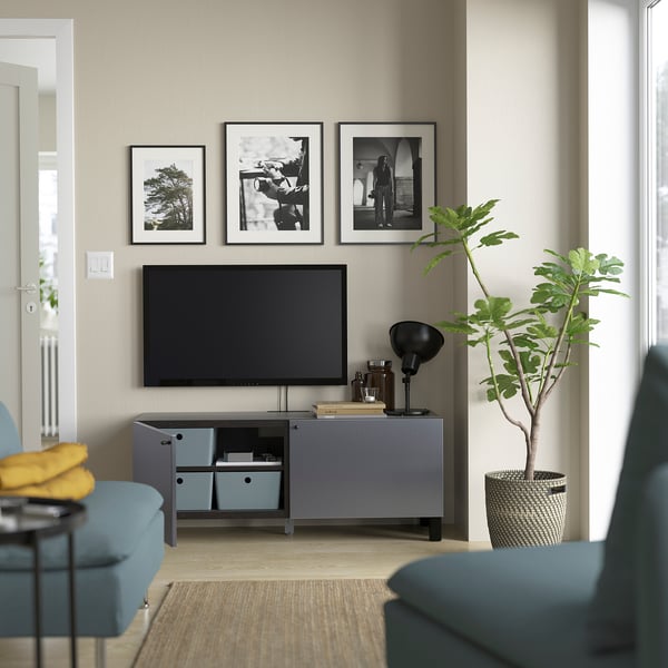 Grey tv stand with blue accents, black lamp, green plant. Tv, framed pictures above. Yellow pillow on blue chair, beige rug.