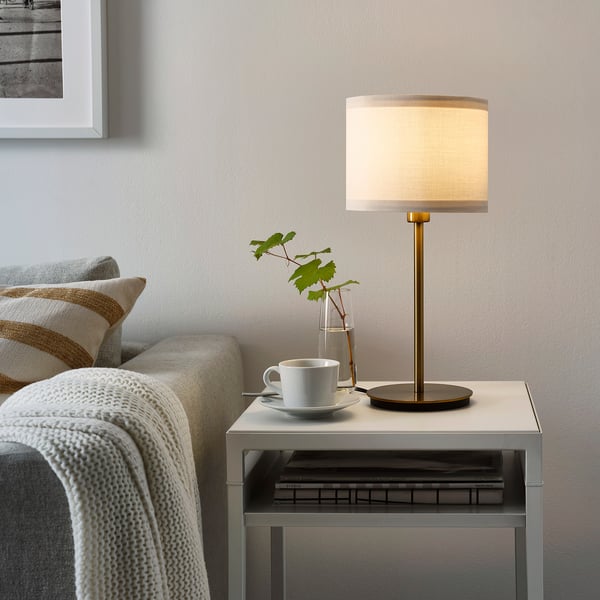 Minimalist RINGSTA lamp with brass base and white shade on side table, in cozy living room setting with couch, blanket, and coffee.
