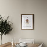A framed picture featuring cake art hangs above a wooden table with a cup, saucer, and carafe.