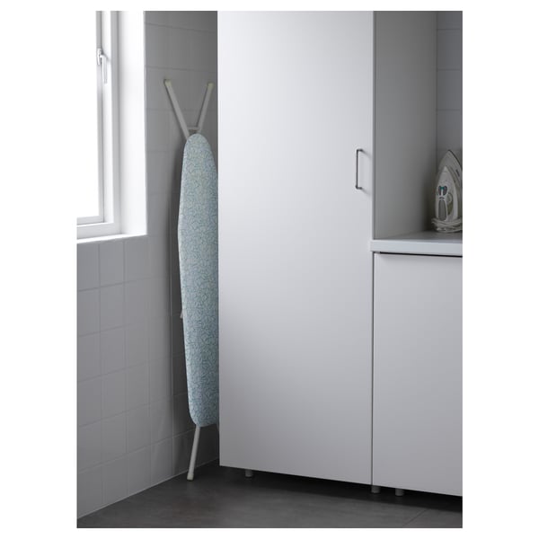 An ironing board set up in a corner, made of steel, with adjustable height. A steam generator is visible.