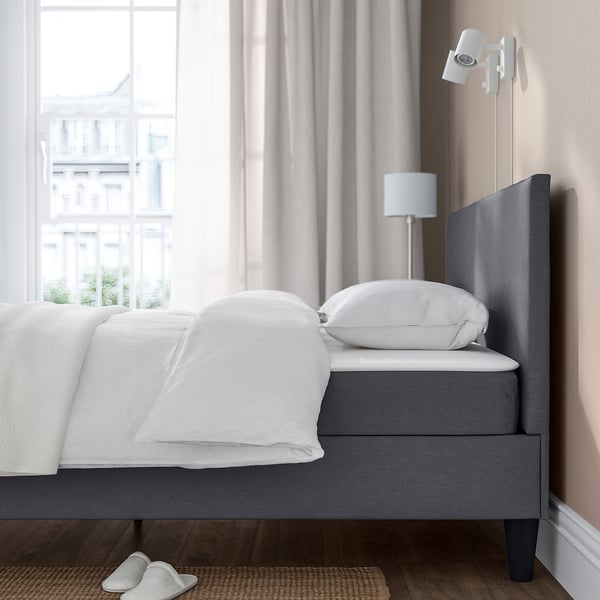 Bed with grey upholstered headboard, white linens, wall lamp, and slippers on a mat, situated by a large window.