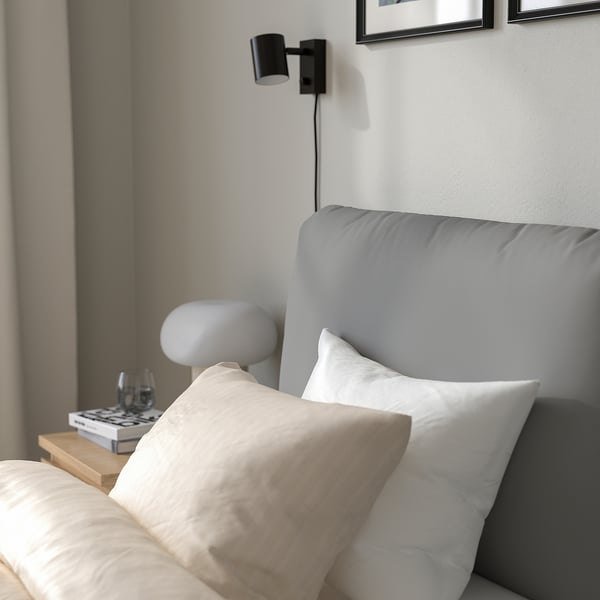 Minimalist bedroom with grey bed, white/beige pillows, side table with lamp, glass, book, wall-mounted reading lamp, soft headboard pillow.