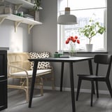 Modern dining with wooden table, black chairs, patterned bench, rattan, leather, and pendant light. Kitchen essentials on shelves.