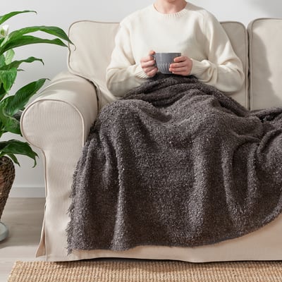 Person sitting on a beige couch holding a mug, covered with a cozy, grey, fringed SANDBRODD throw blanket.