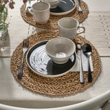 Rustic table setting with gray plates, bowls, and mugs on woven placemats, featuring cutlery, ready for dining.