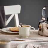 A white ceramic mug filled with coffee sits on a beige tablecloth near a metallic kettle and a small plate with toast.