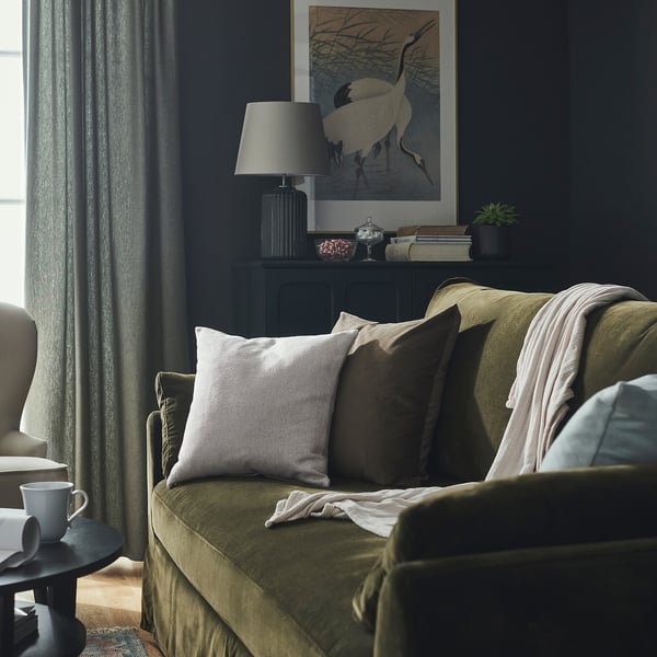 Olive couch with grey pillows, black side table with mug, dark curtains, lamp, crane art.
