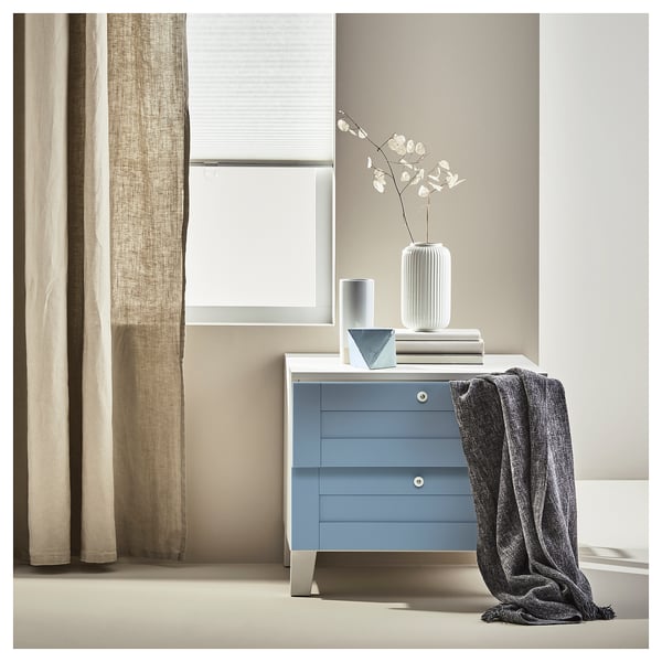 Blue SANNIDAL dresser with grey accents, blanket, vase of white flowers, three drawers, beige curtains.