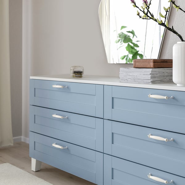Blue dresser with white handles, folded clothes, jar, magazines, mirror, plant.