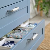Blue chest of drawers, partly open, displaying neatly folded colorful clothes in compartments. Modern handles, organized storage.