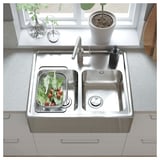 Modern kitchen with stainless steel double sink, fresh veggies in strainer, white countertop, and potted plant.