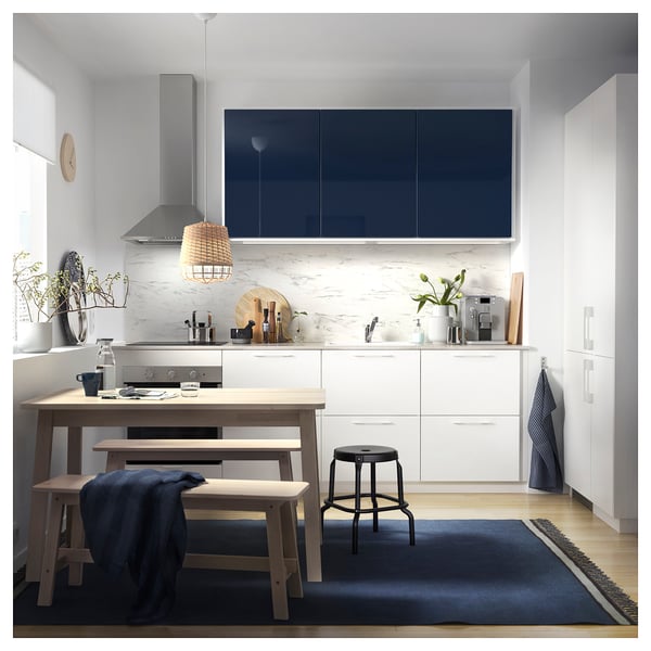 Modern kitchen with blue cabinets, white marble backsplash, wooden table, and black stool. Blue rug and plants decorate the area.