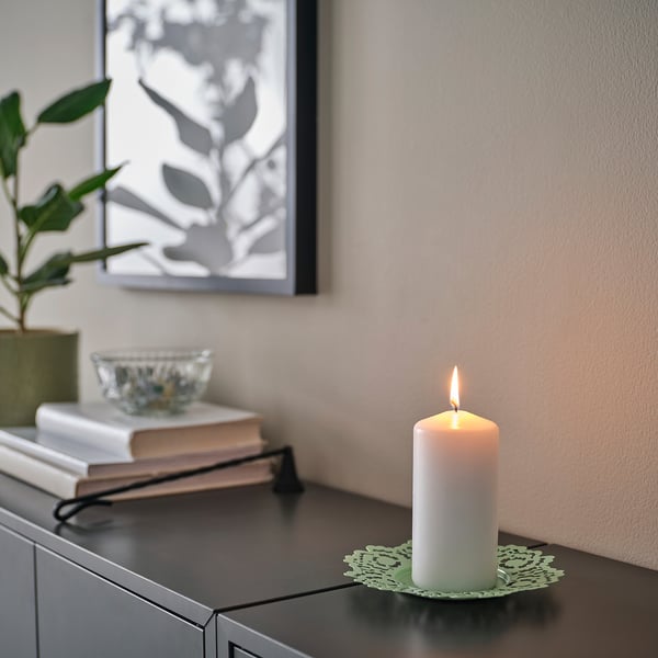 White candle on decorative green dish atop dark surface, beside books and plant.