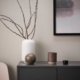 SILKESTRÄD beige candle in ceramic jar on dark shelf with decorative vase and branches.