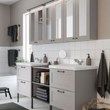Modern grey bathroom with dual sink, mirror, glass storage, towels, and decor.