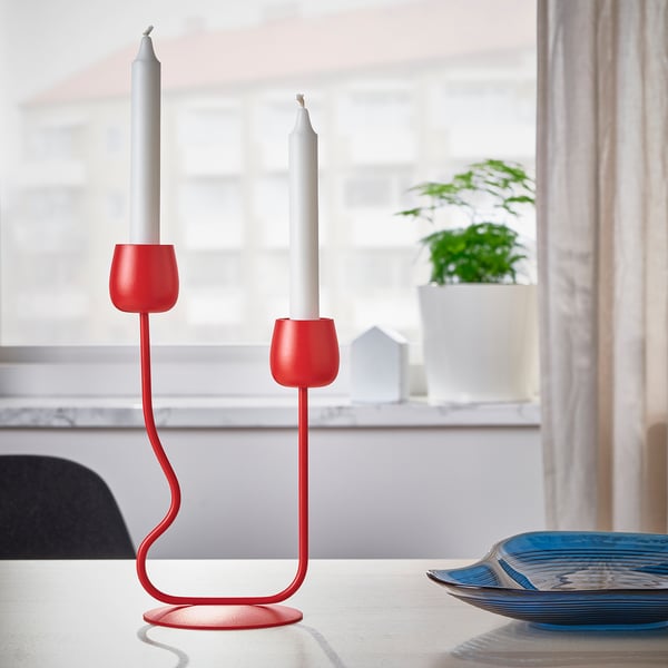 Red candlestick with two white candles on a table, near a blue plate and a plant.