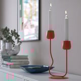 Red candlestick with two lit candles on a shelf, next to a plant and books.