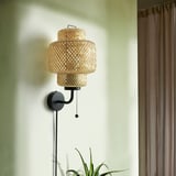Wall-mounted SINNERLIG lamp with woven bamboo shade, dark base, and pull cord. Small, natural, and earthy, sits beside a plant on a white wall.