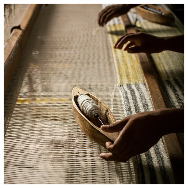 Close-up: hands weaving SJÖTÅTEL rug on loom. Rug: grey, yellow stripes, wool, cotton. Fringed sides.