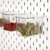 Translucent plastic storage boxes hang on a white pegboard, holding dried herbs and spices. Ideal for kitchen organization and easy access.