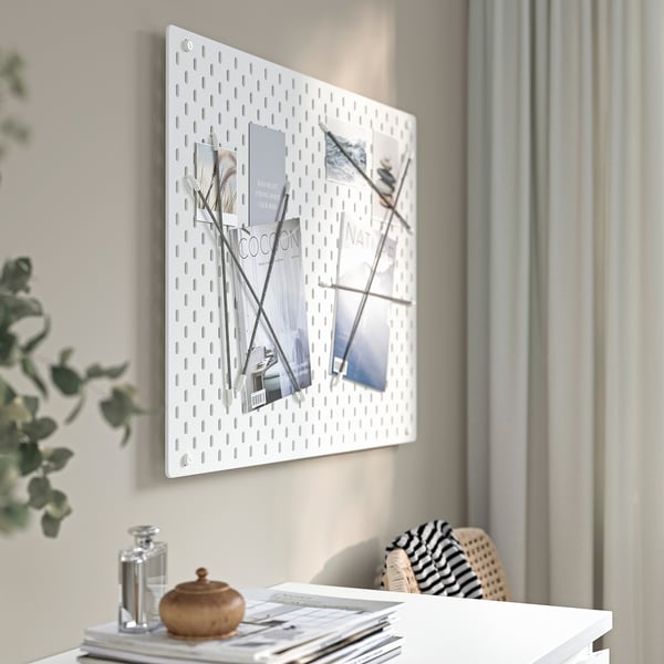 A white wall-mounted SKÅDIS pegboard displays organized photos and magazines. A stylish desk below holds a decorative plant, a jar, and a woven bag.