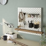 SKÅDIS pegboard organizer on green wall holds cups, brushes, scissors, containers, and a lamp above desk.