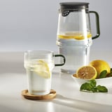 Glass mug with handle and pitcher, lemons, ice, mint; refreshing drink setup.