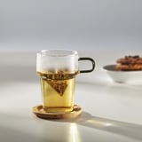 Clear glass mug with green handle holds a tea bag, placed on wooden coaster, sitting on table with sunlight reflecting off it.