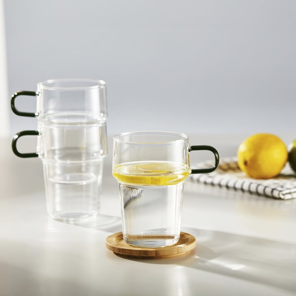 Two stackable glasses, one with lemon slice in water, on wooden coaster, against white background.