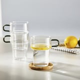 Two stackable glasses, one with lemon slice in water, on wooden coaster, against white background.