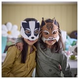 Two kids in badger & lynx masks smile in cozy room.