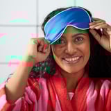 Smiling woman, adjusting blue silk eye mask, wrapped in striped pink robe.