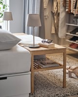 Bedside table with wooden frame and white table lamp with gray shade, beside a bed.