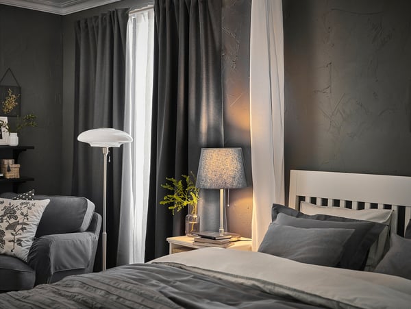 Bedroom with black walls, white bed with gray pillows, gray curtains, gray lamp with SKOTTORP white shade, and gray armchair by window.