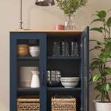 Blue cabinet with glass doors displaying bowls, glasses, and baskets; oak top, greenery beside.
