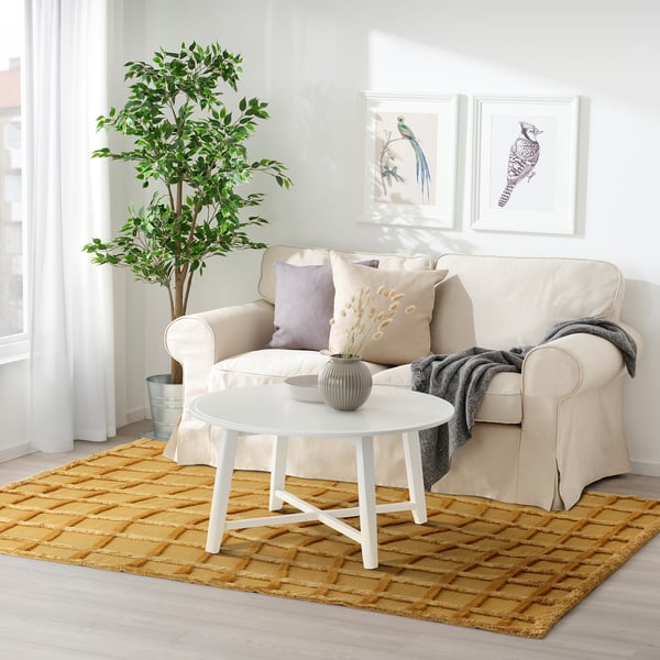Cozy living room with beige sofa, white coffee table, and yellow woven rug. Artwork and plant enhance the decor.