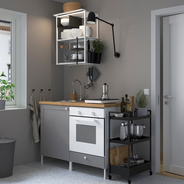 Modern kitchen with grey cabinets and white oven. Open shelves hold plates and plants. Black cart with wheels holds cooking items.