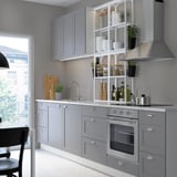 Modern kitchen with gray cabinets and sleek appliances. Open shelves hold dishes and plants. Built-in oven and sink near a large window with chair.