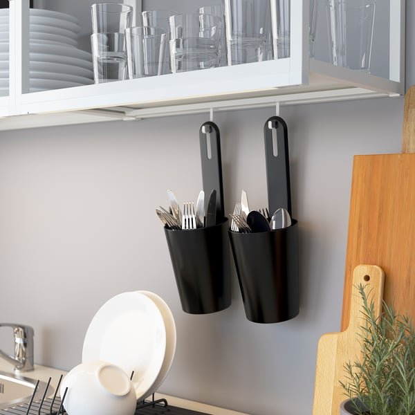 Kitchen counter with utensils: black SKYDRAG containers holding forks, knives, and spoons. Above, a white cabinet with clear glasses and plates.