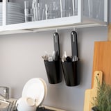 Kitchen counter with utensils: black SKYDRAG containers holding forks, knives, and spoons. Above, a white cabinet with clear glasses and plates.