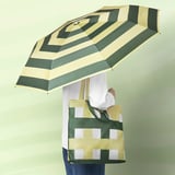 Person holding open umbrella with green and beige stripes, also carrying matching shoulder bag with squares.
