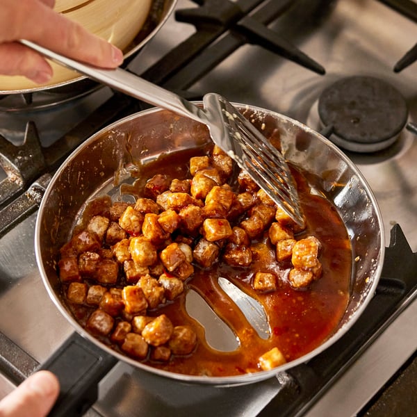 Person cooking with a stainless-steel pan on a gas stove, using metal utensils; the pan has detachable handles.