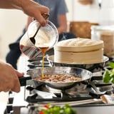 Person pours liquid from a jug into a skillet on a stove next to a steamer.