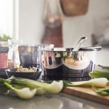 A shiny saucepan with detachable handle, surrounded by veggies and herbs, ready for cooking.