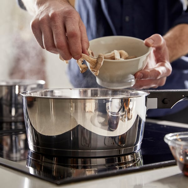 Person attaching handle to stainless-steel saucepan on stove. Removable handle for easy storage, suitable for all hob types.
