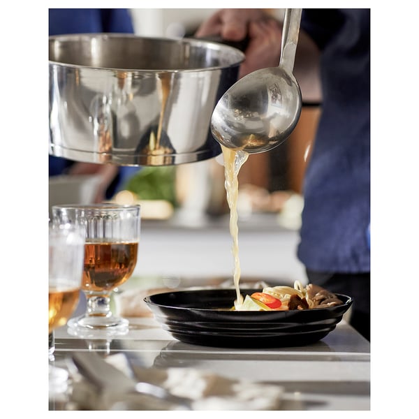 Silver ladle pouring soup into black bowl, next to glass of liquid on white table.