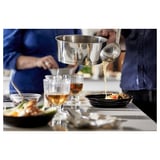 A chef pours sauce over a dish in a kitchen. Glasses and plates are on the table.