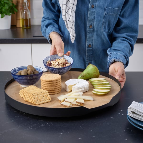 Person arranging snacks on a wooden spinning tray with crackers, cheese, fruit, and nuts on a kitchen counter.