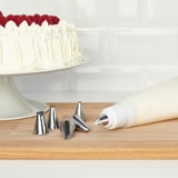 A cake decorating set with piping bag and tips. A frosted cake with berries is nearby.